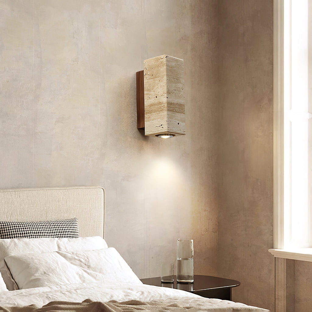 Square-sided travertine Clérachic wall light beside a sunlit window, with white bedding and a glass of water below it.