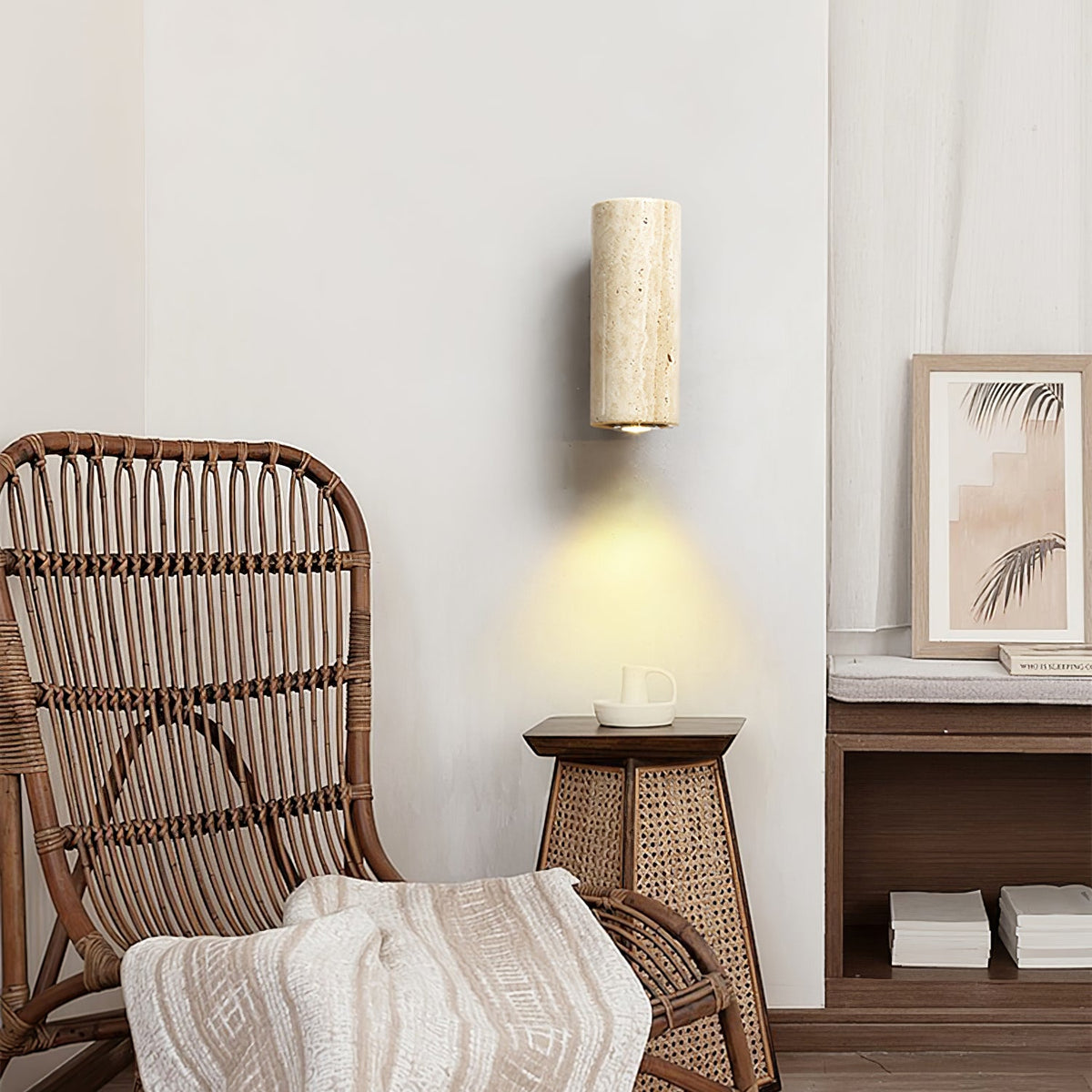 Cylindrical travertine wall light from Clérachic lit above a rattan chair, a woven side table and a framed leaf print.