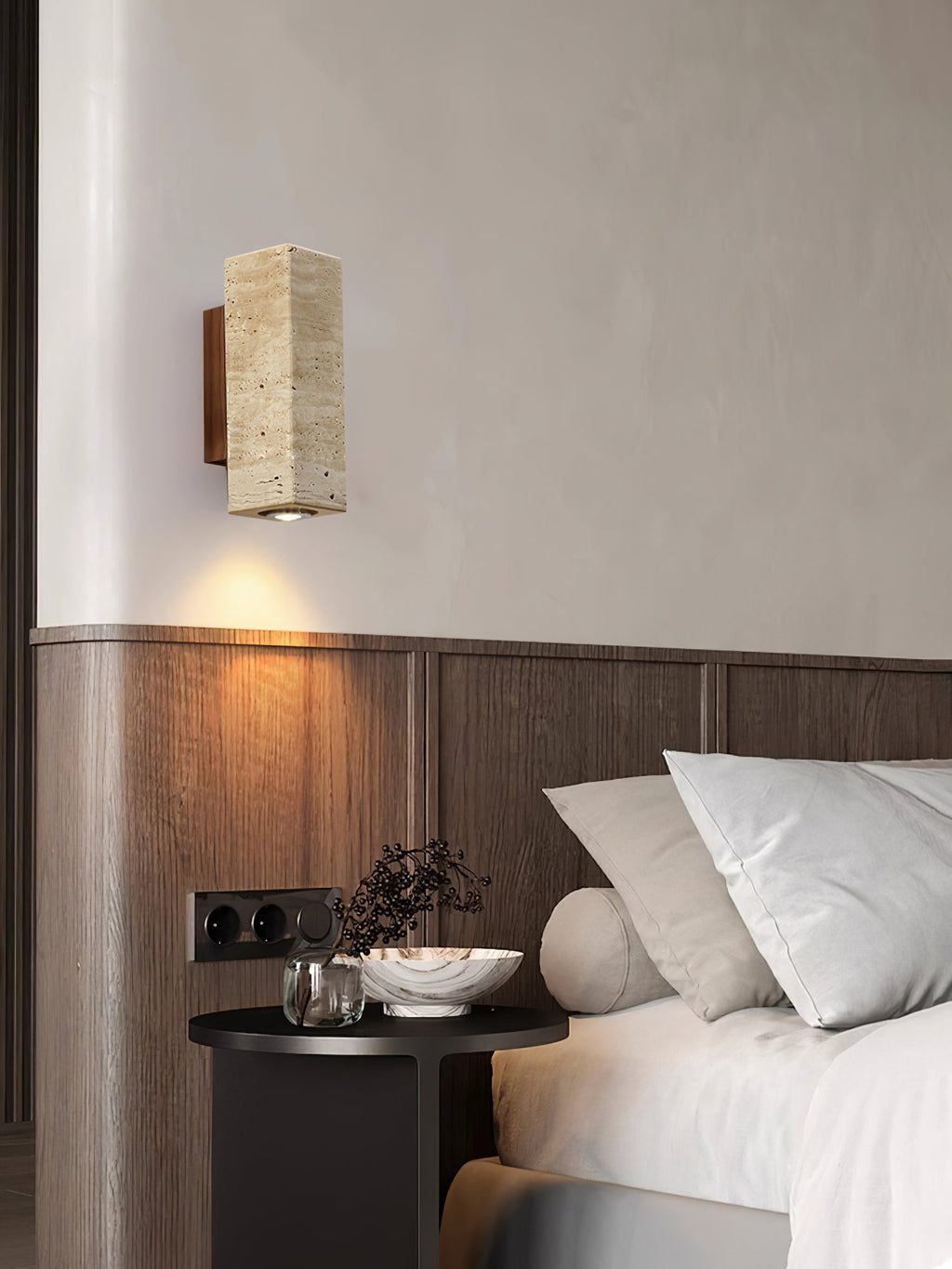 Clérachic travertine wall light with a walnut-colour back, above a dark bedside table and a panelled headboard wall.
