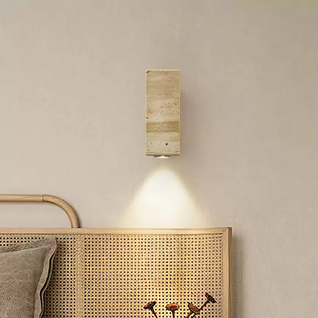 Square-sided travertine wall light from Clérachic, lit, above a rattan headboard on a smooth beige wall.