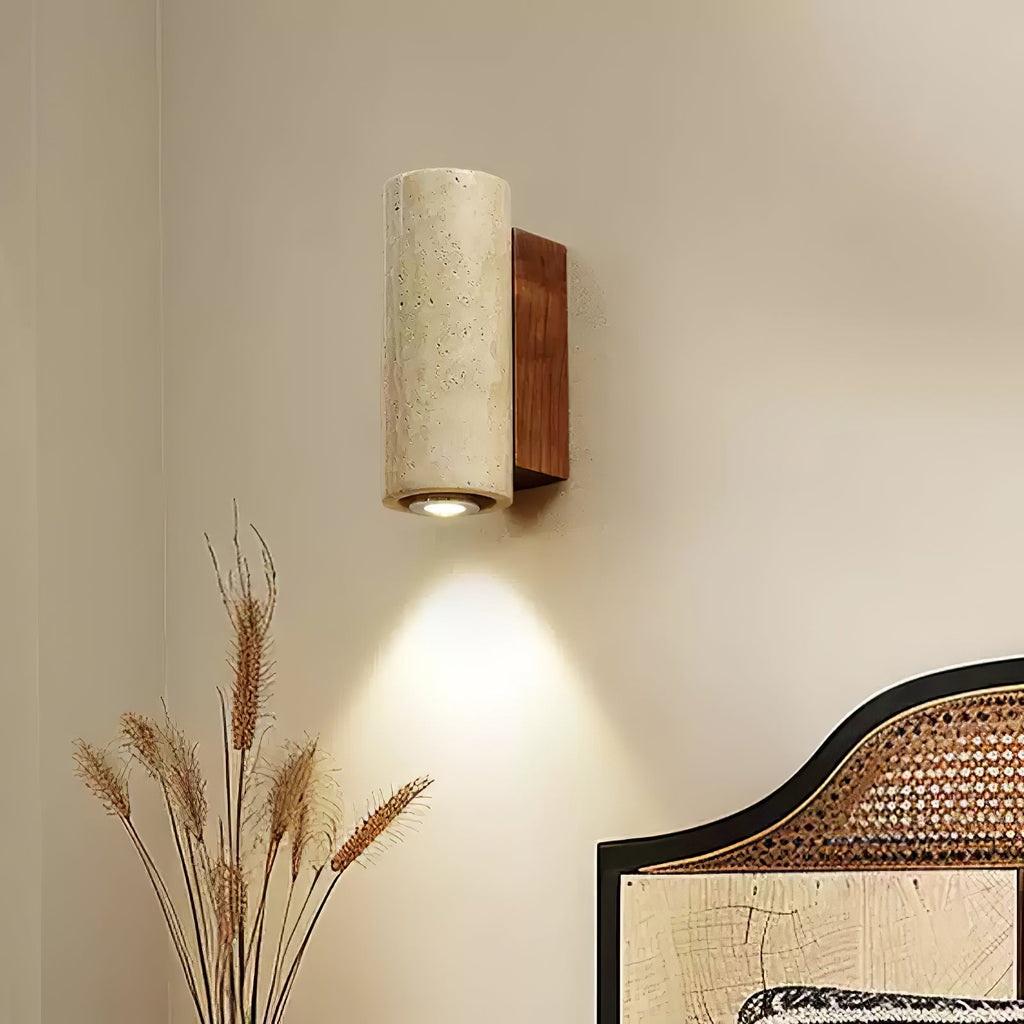 Cylindrical Clérachic travertine wall light with its downlight on, beside dried wheat stems and a rattan bedhead.