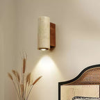 Cylindrical Clérachic travertine wall light with its downlight on, beside dried wheat stems and a rattan bedhead.