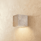 Clérachic cube wall light cut from pale travertine, its natural pitted stone showing, casting warm light downward on plaster.