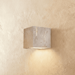 Clérachic cube wall light cut from pale travertine, its natural pitted stone showing, casting warm light downward on plaster.