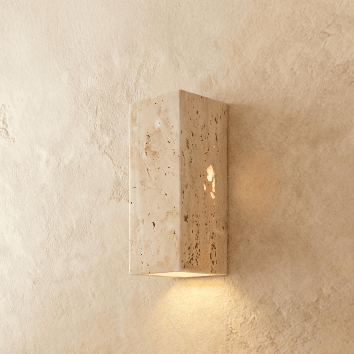 Clérachic rectangular travertine wall light with a pitted, veined stone surface, glowing warm from its base onto the wall.