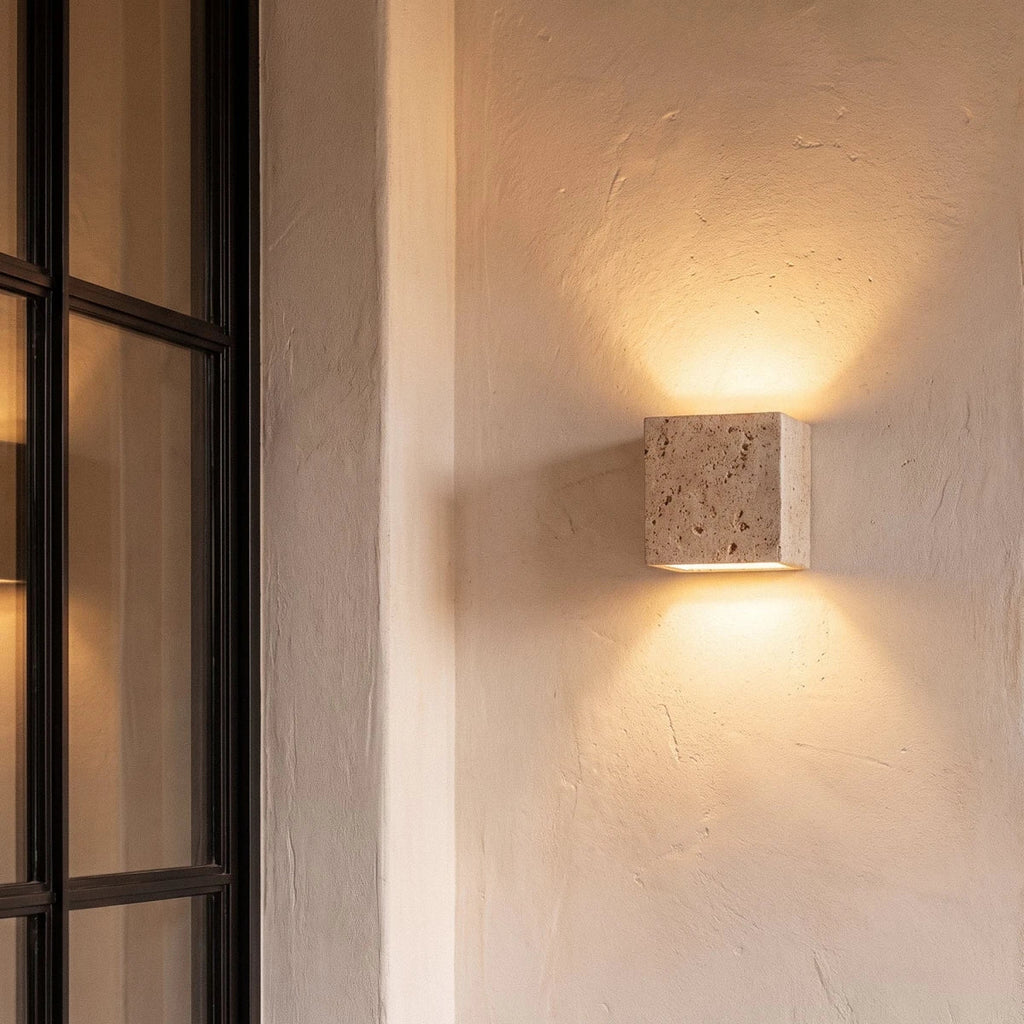 Clérachic travertine cube wall light on a plaster wall beside a black-framed door, casting warm light up and down.