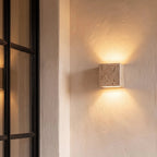Clérachic travertine cube wall light on a plaster wall beside a black-framed door, casting warm light up and down.