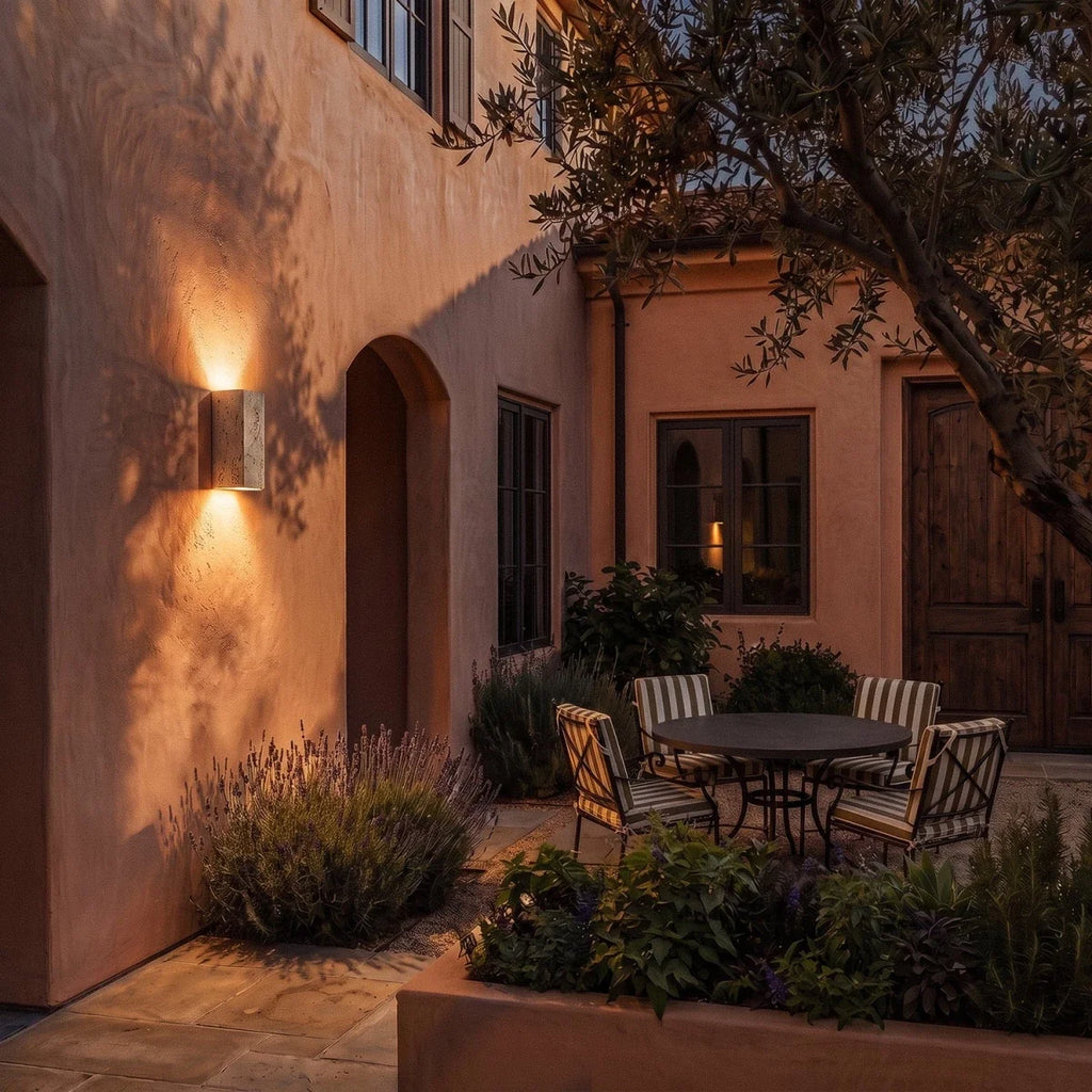 Clérachic tall travertine wall light glowing warm at dusk on a terracotta courtyard wall above lavender and a bistro table.