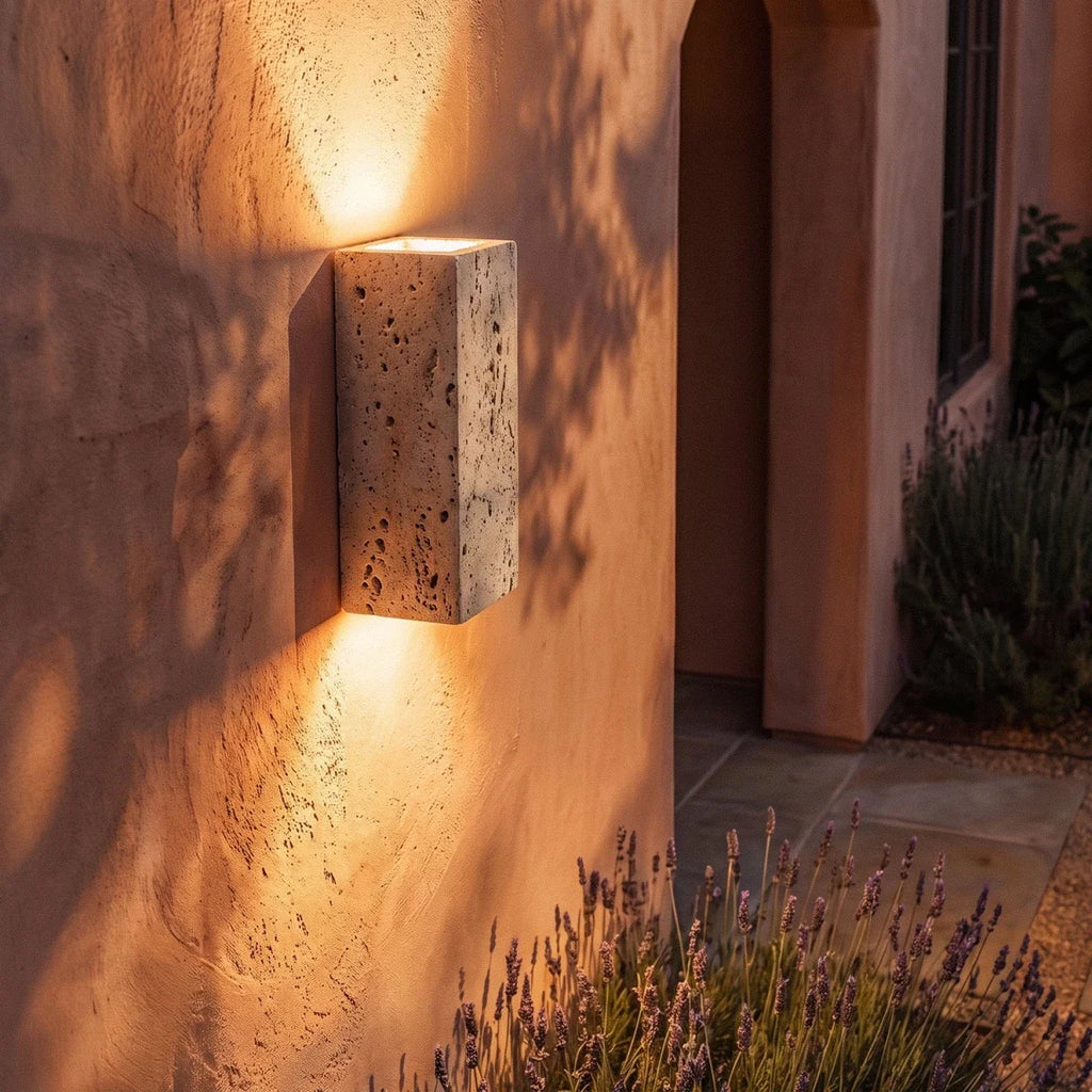 Clérachic rectangular travertine wall light close up, its pitted stone face lit above and below on a warm plaster wall.