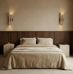 A pair of Clérachic travertine wall lights flank a bed with a dark wood headboard panel and stone bedside tables.