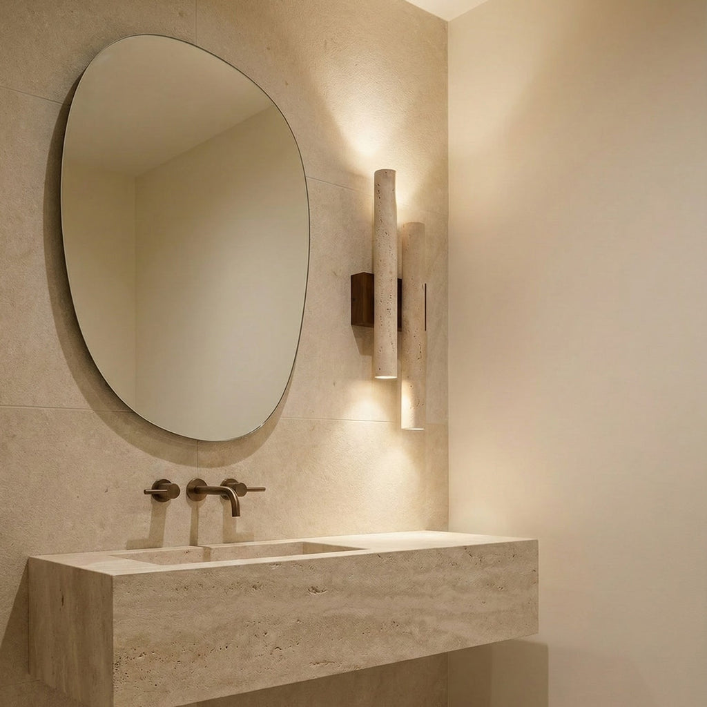 A Clérachic wall light glows beside an oval mirror in a bathroom with a travertine basin and a wall-mounted brass tap.