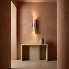 A Clérachic travertine wall light lights a terracotta-toned alcove, above a stone console with a dry branch in a vase.