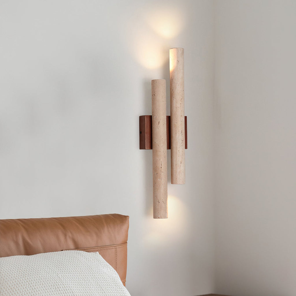 Lit Clérachic wall light with two stone tubes and a walnut backplate, on a pale wall above a tan leather headboard.