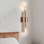 Lit Clérachic wall light with two stone tubes and a walnut backplate, on a pale wall above a tan leather headboard.