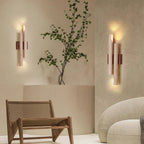 Two Clérachic travertine wall lights on either side of a corner with a tall branch in a vase and a woven wooden chair.