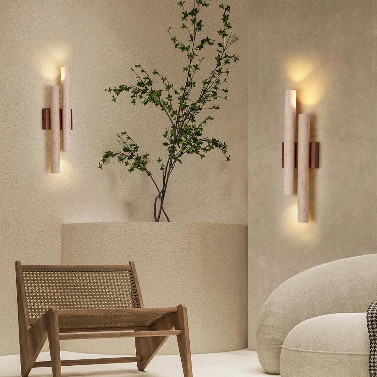 Two Clérachic travertine wall lights on either side of a corner with a tall branch in a vase and a woven wooden chair.
