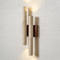 Close view of this Clérachic wall light with two travertine tubes and a walnut-wood plate, lit on a pale textured wall.