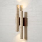 Close view of this Clérachic wall light with two travertine tubes and a walnut-wood plate, lit on a pale textured wall.