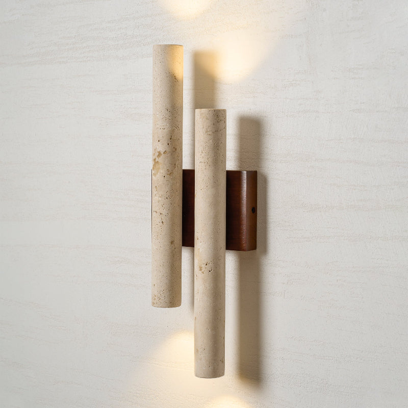 Close view of this Clérachic wall light with two travertine tubes and a walnut-wood plate, lit on a pale textured wall.