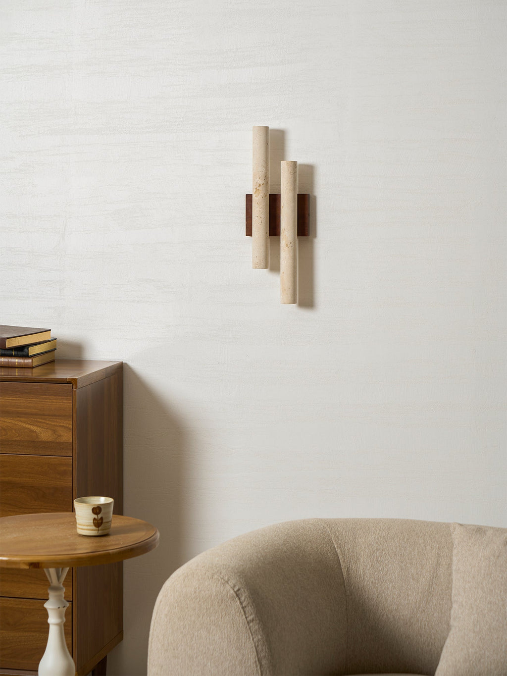Unlit Clérachic wall light on a white wall, above a wooden chest of drawers, a small table with a cup and a beige armchair.