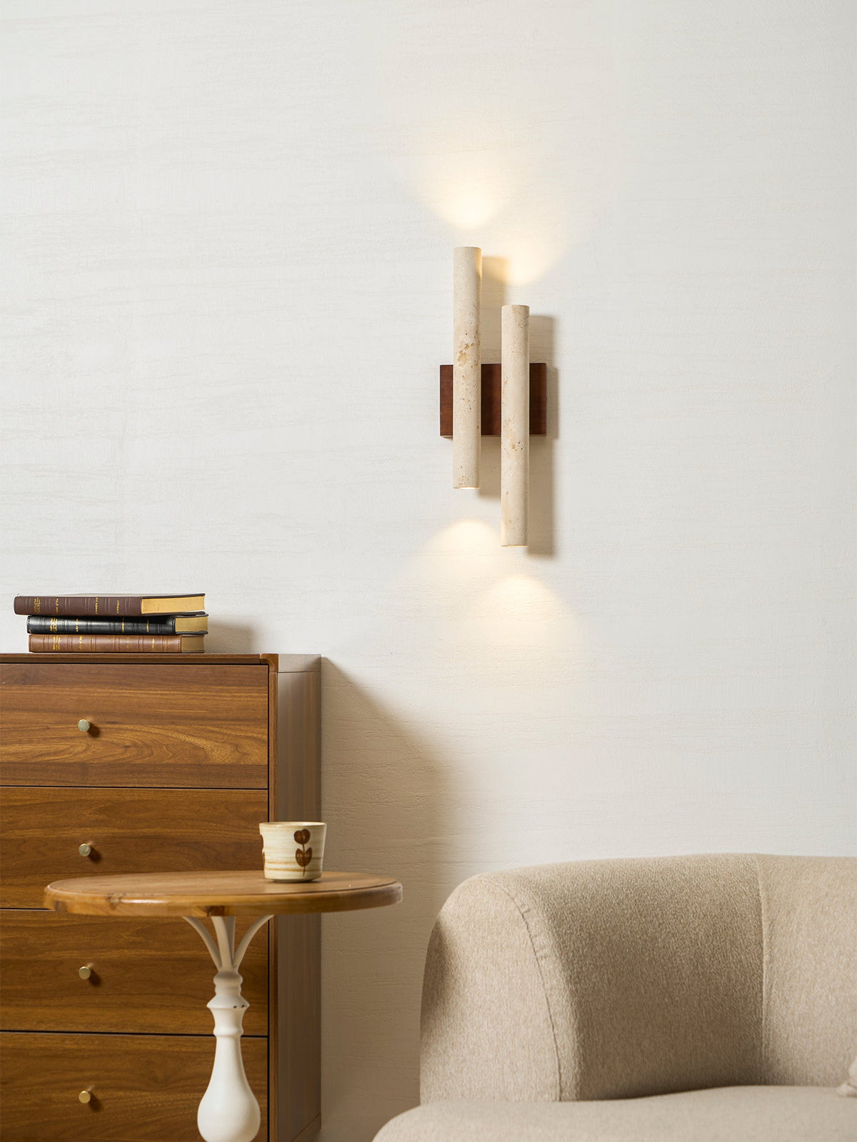 The Clérachic wall light lit, casting a pool of light up and down a white wall beside a chest of drawers and a side table.