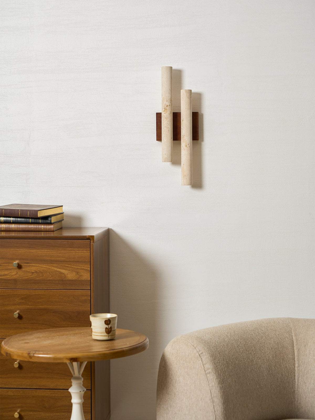 An unlit Clérachic wall light above a wooden chest and a round side table, with a beige chair at the corner of the frame.