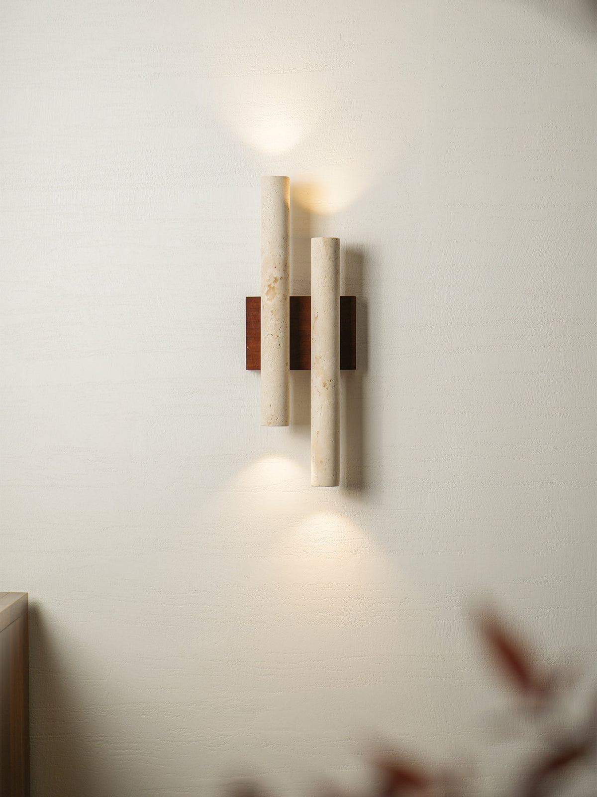 Soft light spills up and down from this Clérachic travertine wall light, with a blurred dark leaf in the corner.