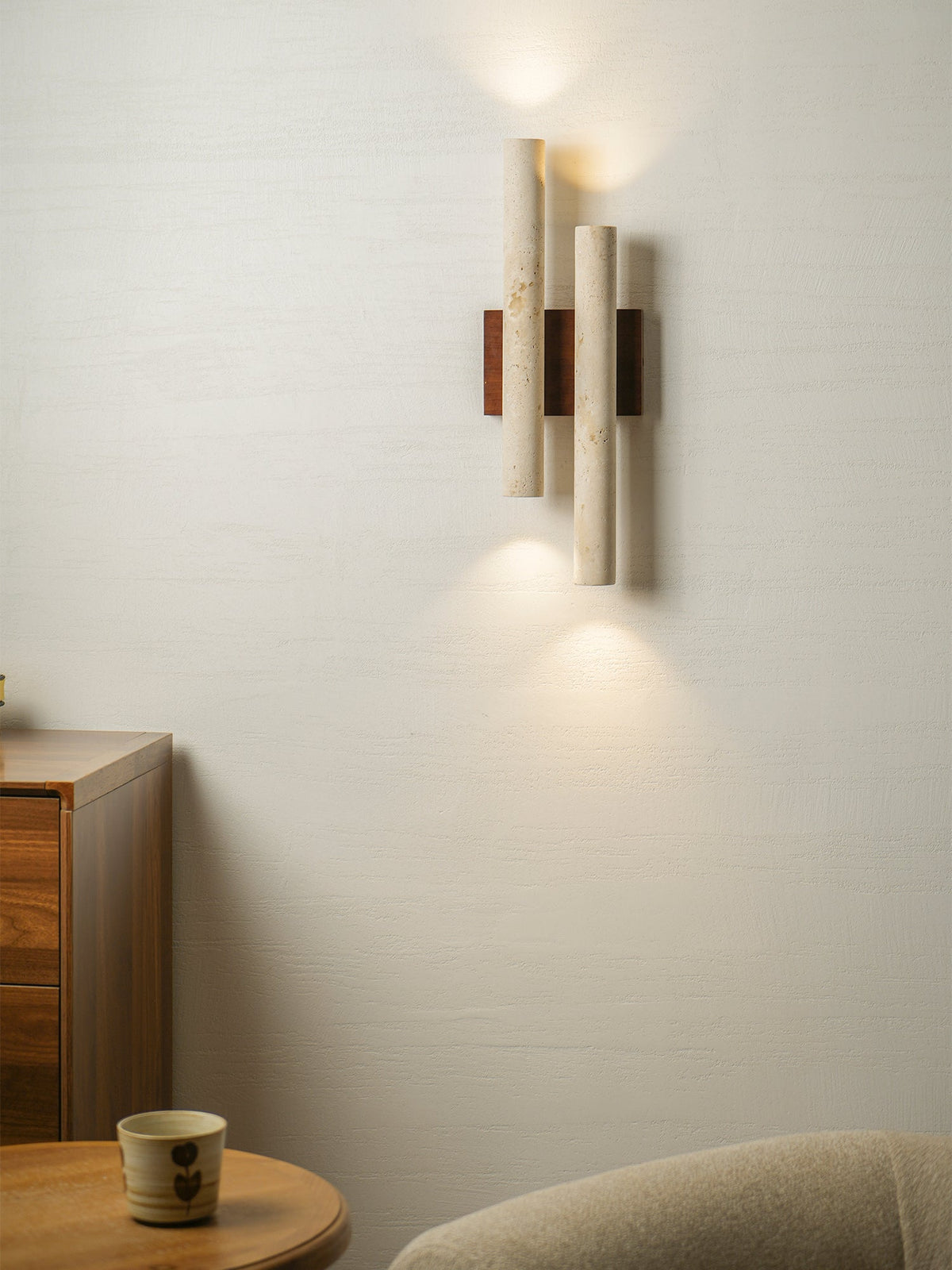 A Clérachic wall light with three-quarter view of its stone tubes lit on a pale wall, beside a wooden drawer unit.