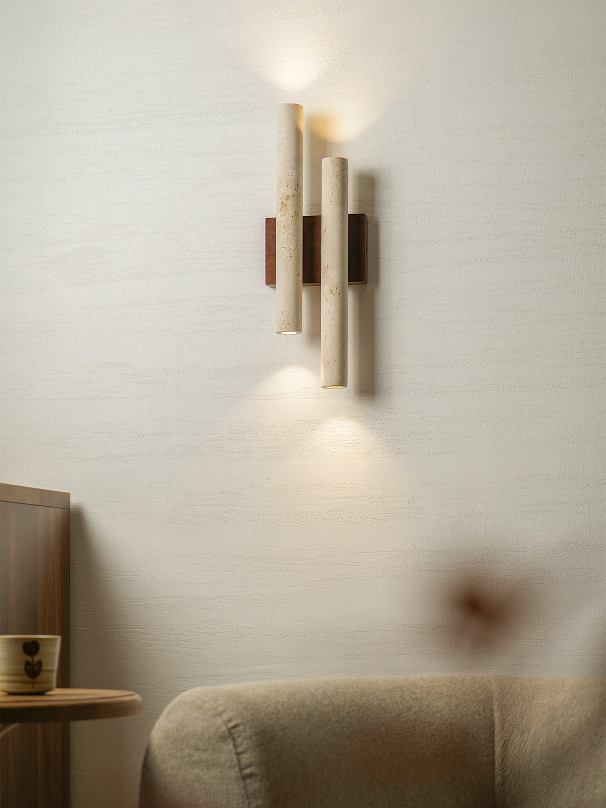 This Clérachic travertine wall light glows on a white wall, with a side table and a beige armchair in the foreground.