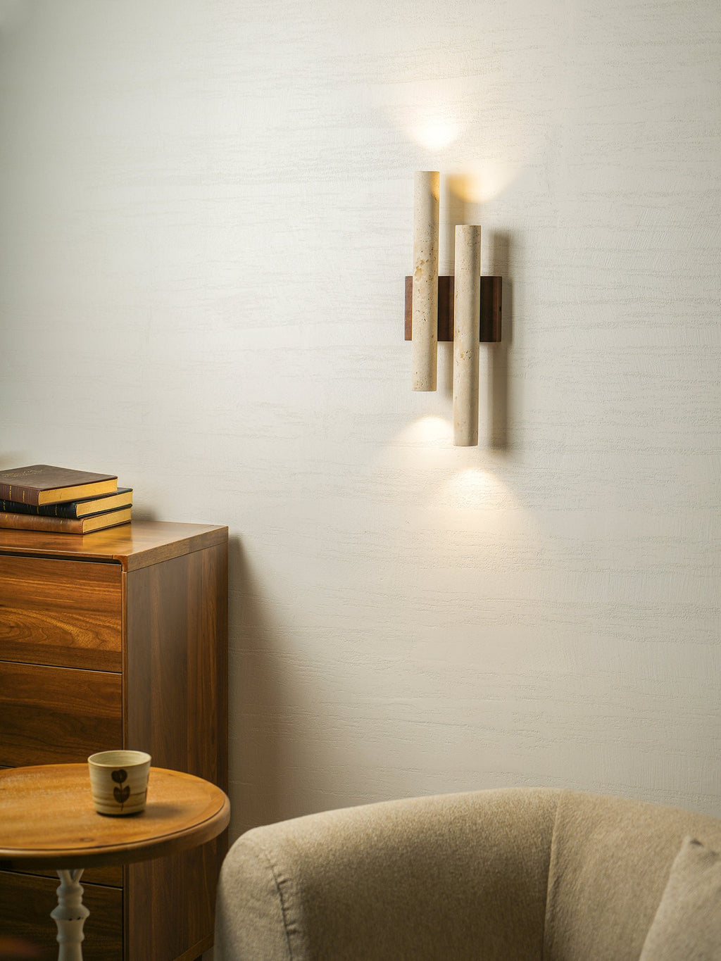 A Clérachic travertine wall light glows beside a wooden chest and a round table, with a beige chair beneath.