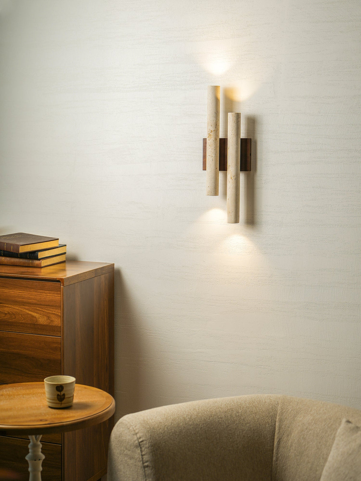 A Clérachic travertine wall light glows beside a wooden chest and a round table, with a beige chair beneath.