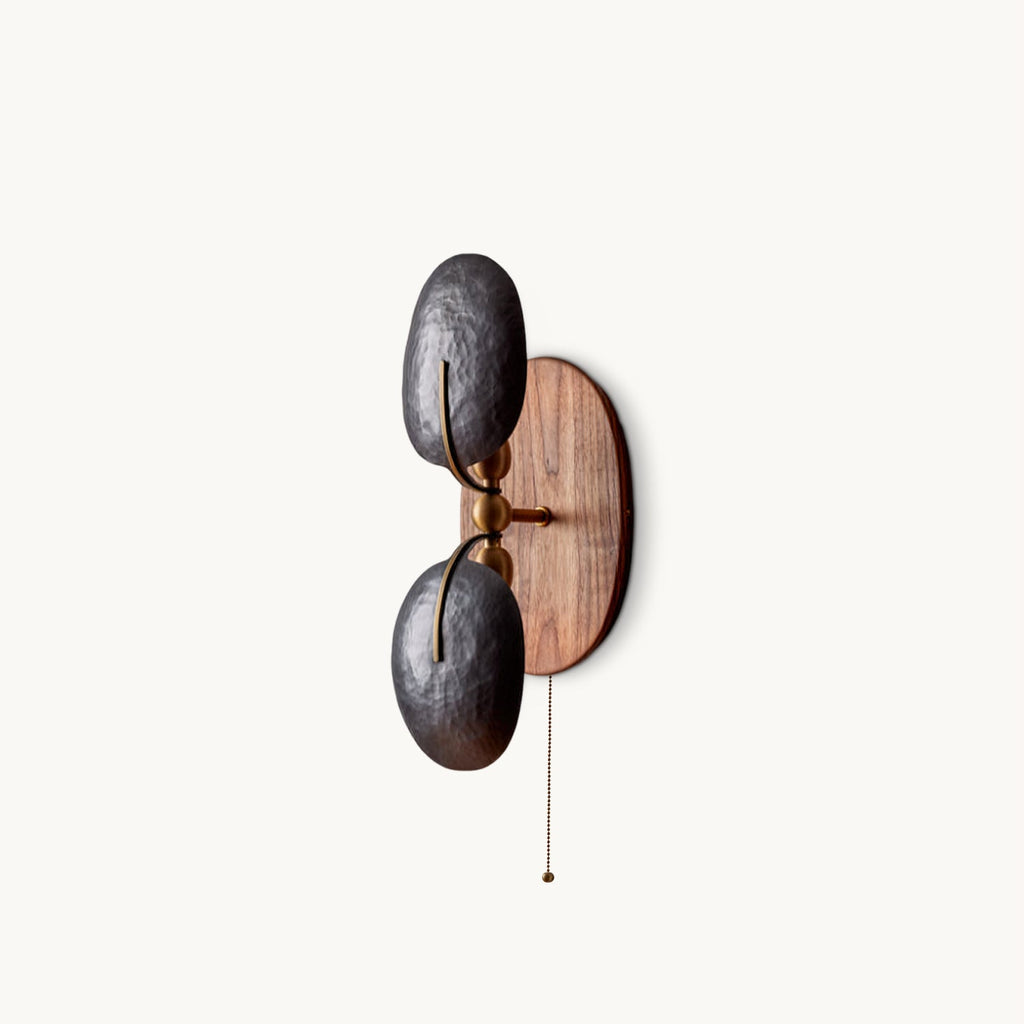 A Clérachic twin-shade wall light with two hammered dark metal shades on a walnut backplate and a pull chain, on a plain white backdrop.
