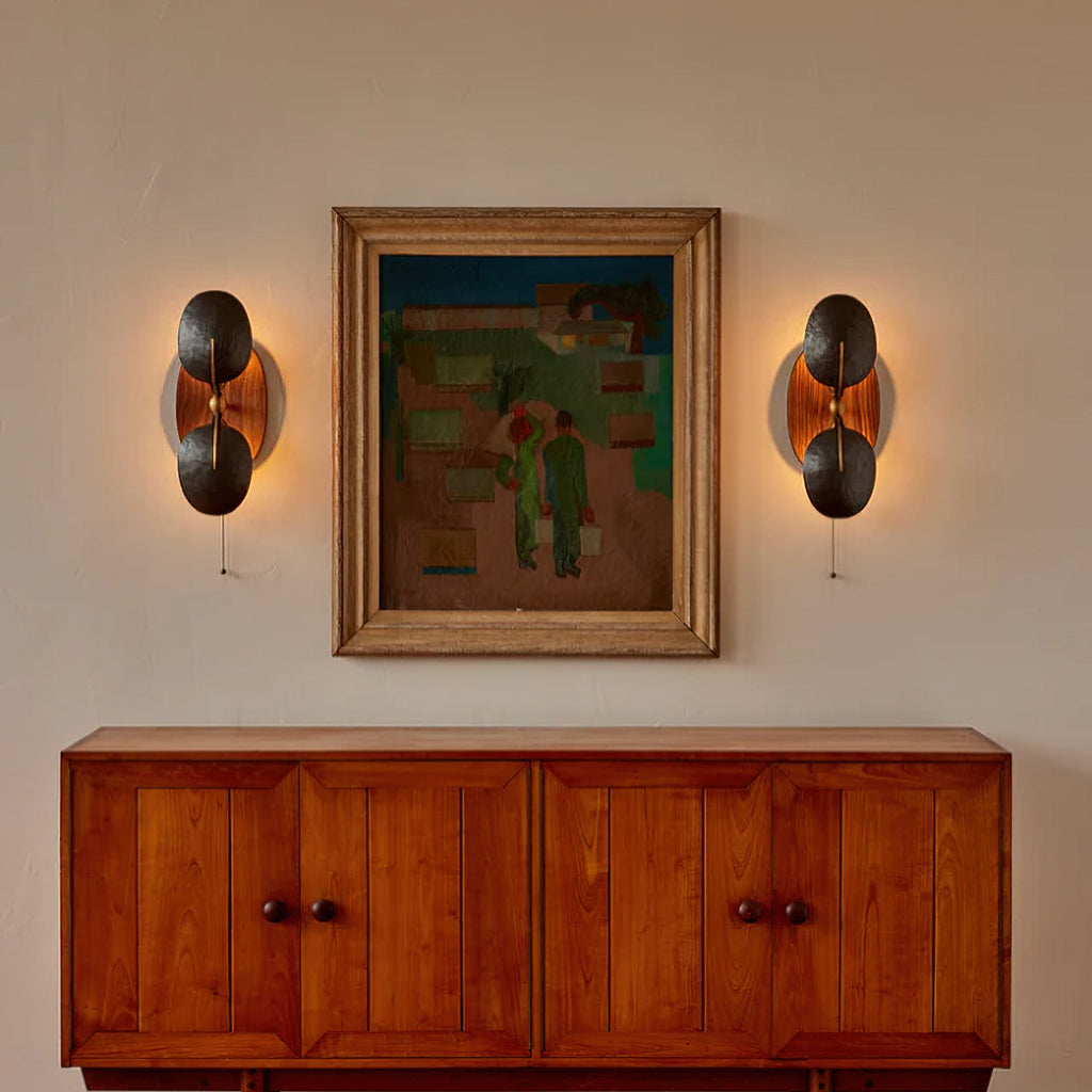 A pair of Clérachic wall lights glowing either side of a framed painting, above a teak-toned sideboard with panelled doors.