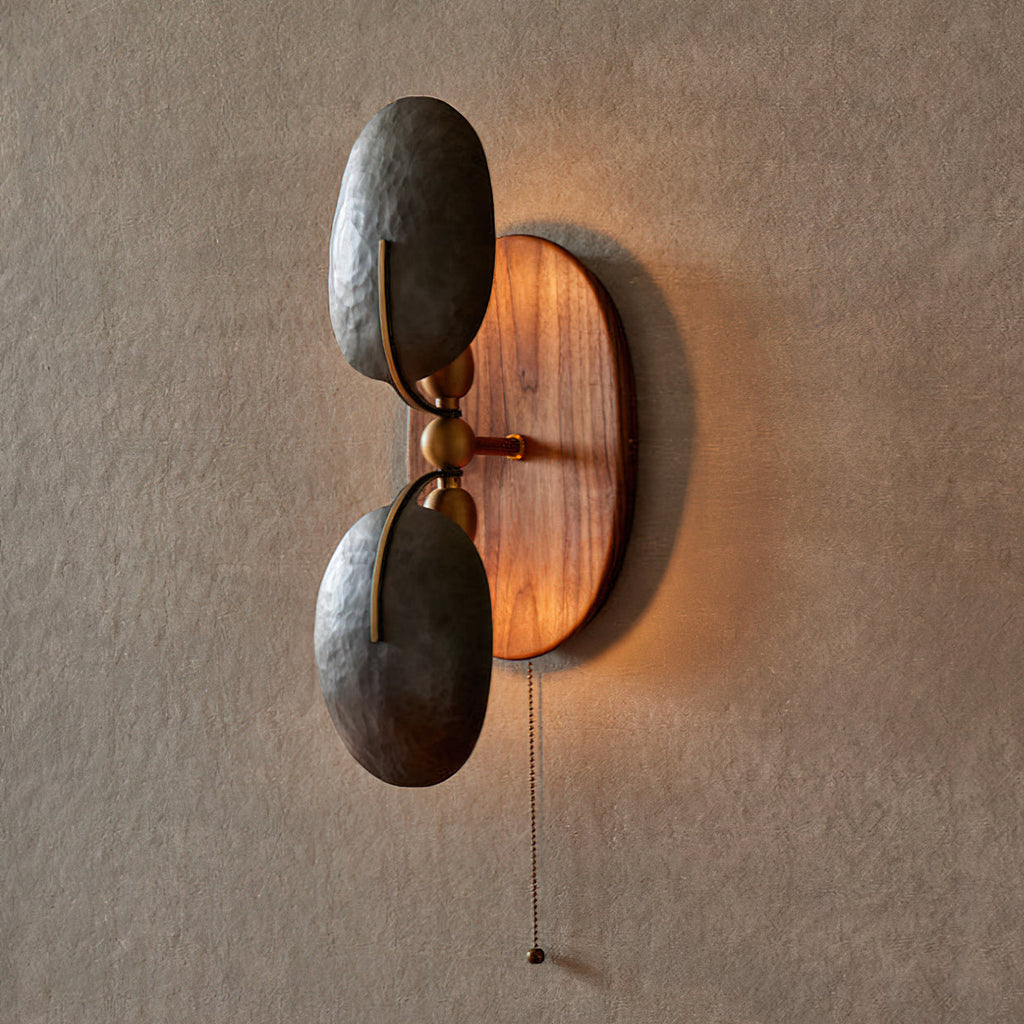 Lit Clérachic wall light on a textured grey wall, with warm light glowing around the oval walnut backplate behind two dark shades.