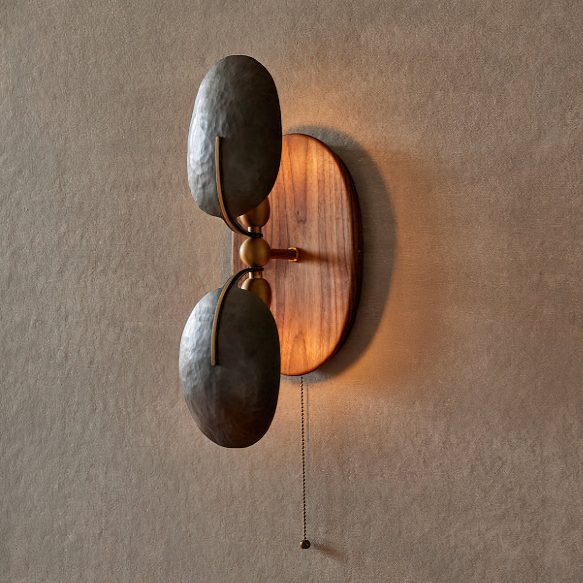 Lit Clérachic wall light on a textured grey wall, with warm light glowing around the oval walnut backplate behind two dark shades.