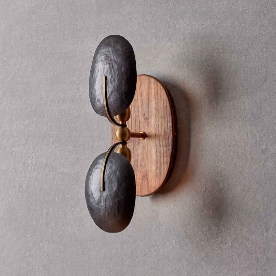 This Clérachic wall light unlit on a grey wall, showing two hammered dark shades, brass joints and an oval walnut backplate.