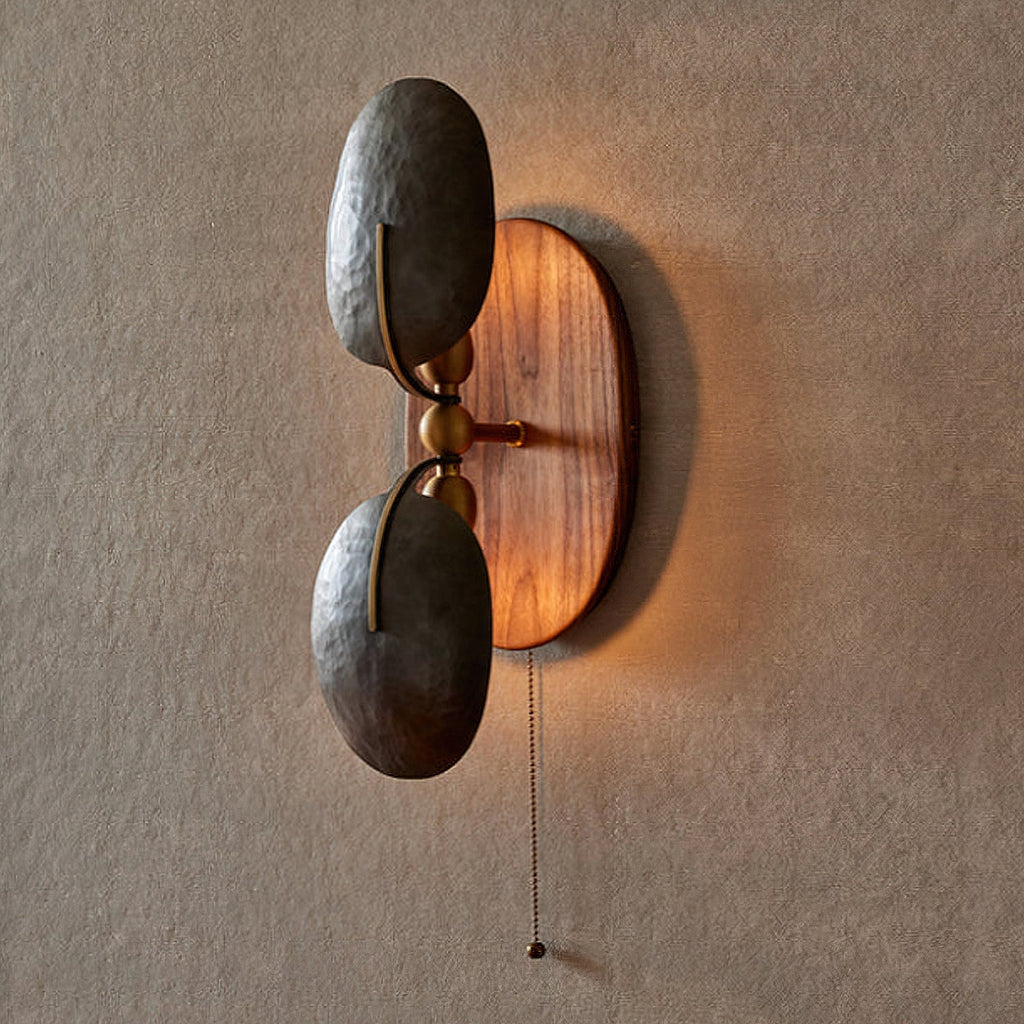 A lit Clérachic wall light with a pull chain, its walnut backplate glowing softly behind two dark hammered shades on a grey wall.