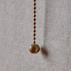 Close view of the beaded pull chain and round brass pull on this Clérachic wall light, hanging against a woven grey wall.