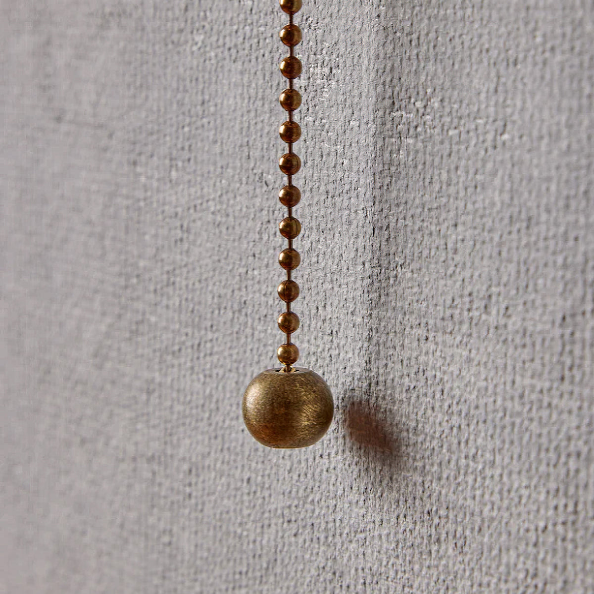 Close view of the beaded pull chain and round brass pull on this Clérachic wall light, hanging against a woven grey wall.
