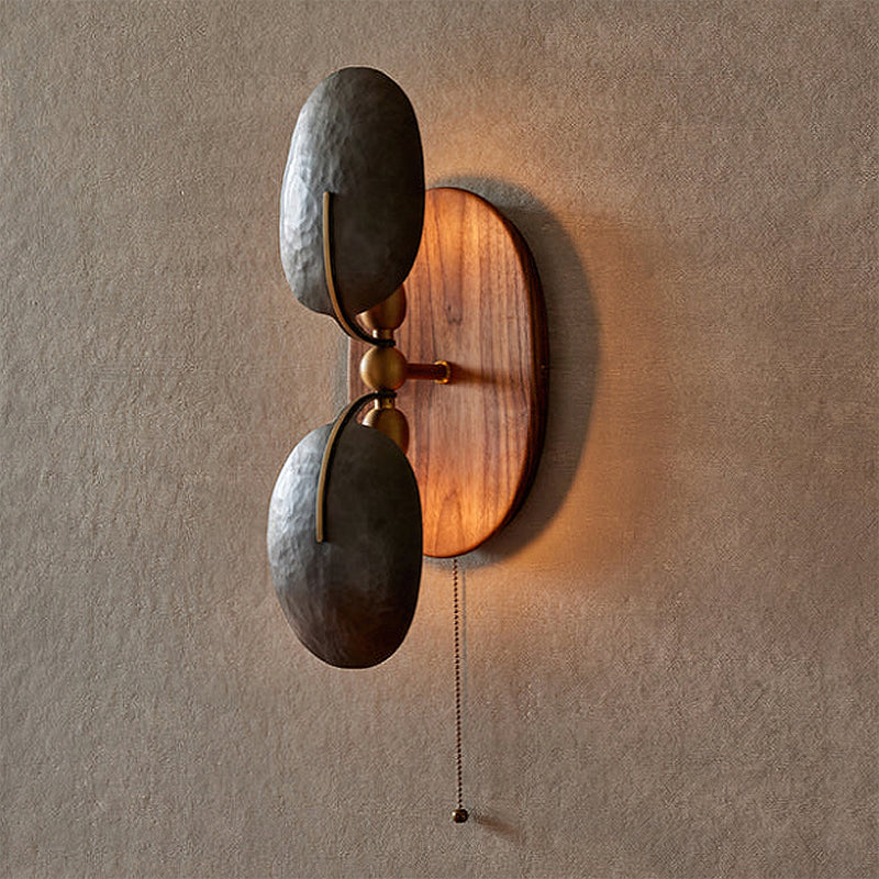 Warm light glows from behind the oval walnut backplate of this Clérachic wall light, with two dark shades and a pull chain below.