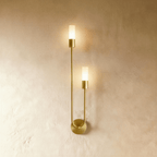 Clérachic brass wall light with two slim frosted cylinder diffusers on a U-shaped arm and round wall plate, lit warm on plaster.