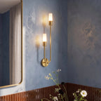 Clérachic brass wall light close-up, two frosted cylinders glowing on a blue plaster wall beside a brass-framed mirror.