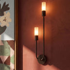 Clérachic black wall light close-up, two glowing frosted cylinder diffusers on a slim U-shaped arm against a terracotta wall.