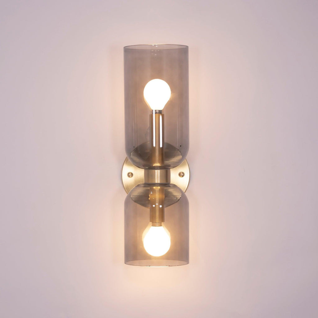 In gold, this Clérachic wall light holds smoke grey glass cylinders with the bulbs visible.