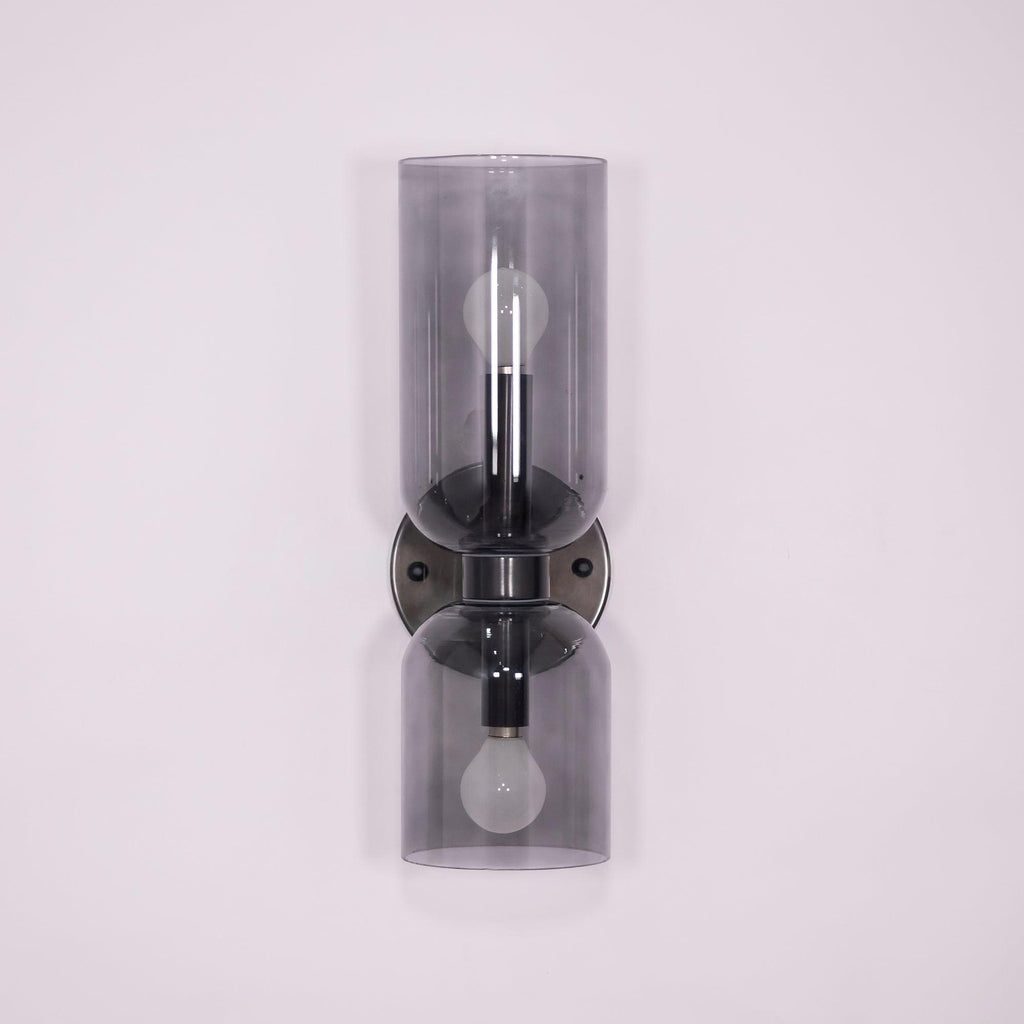 A gunmetal Clérachic sconce holds smoke grey glass cylinders, unlit against a lilac backdrop.