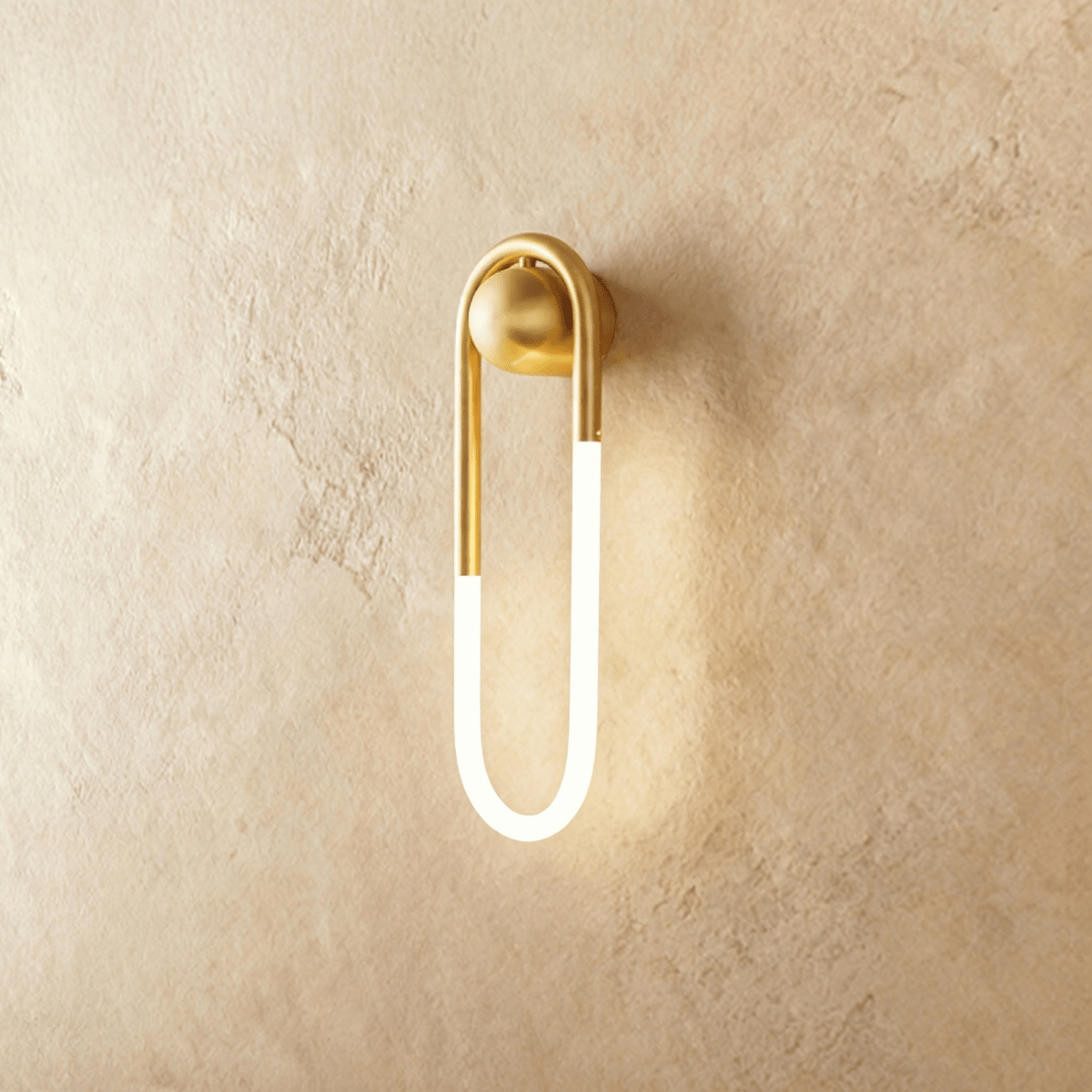 Clérachic elongated U-shaped wall light in brushed brass with a frosted glowing tube, mounted on warm limewash plaster.