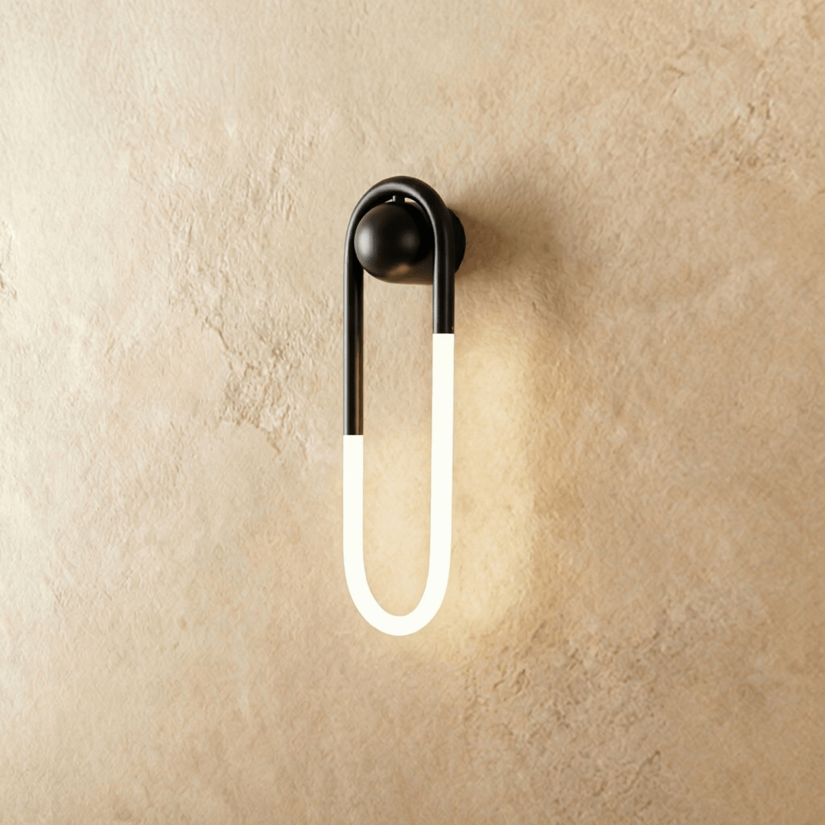 Clérachic elongated U-shaped wall light with a matt black arch and ball mount and a glowing frosted tube on limewash plaster.