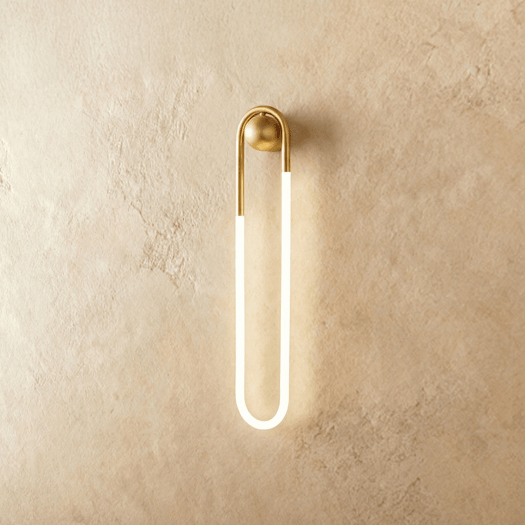 Clérachic slim U-shaped wall light with a brass arch and ball mount and a softly glowing frosted tube, on limewash plaster.