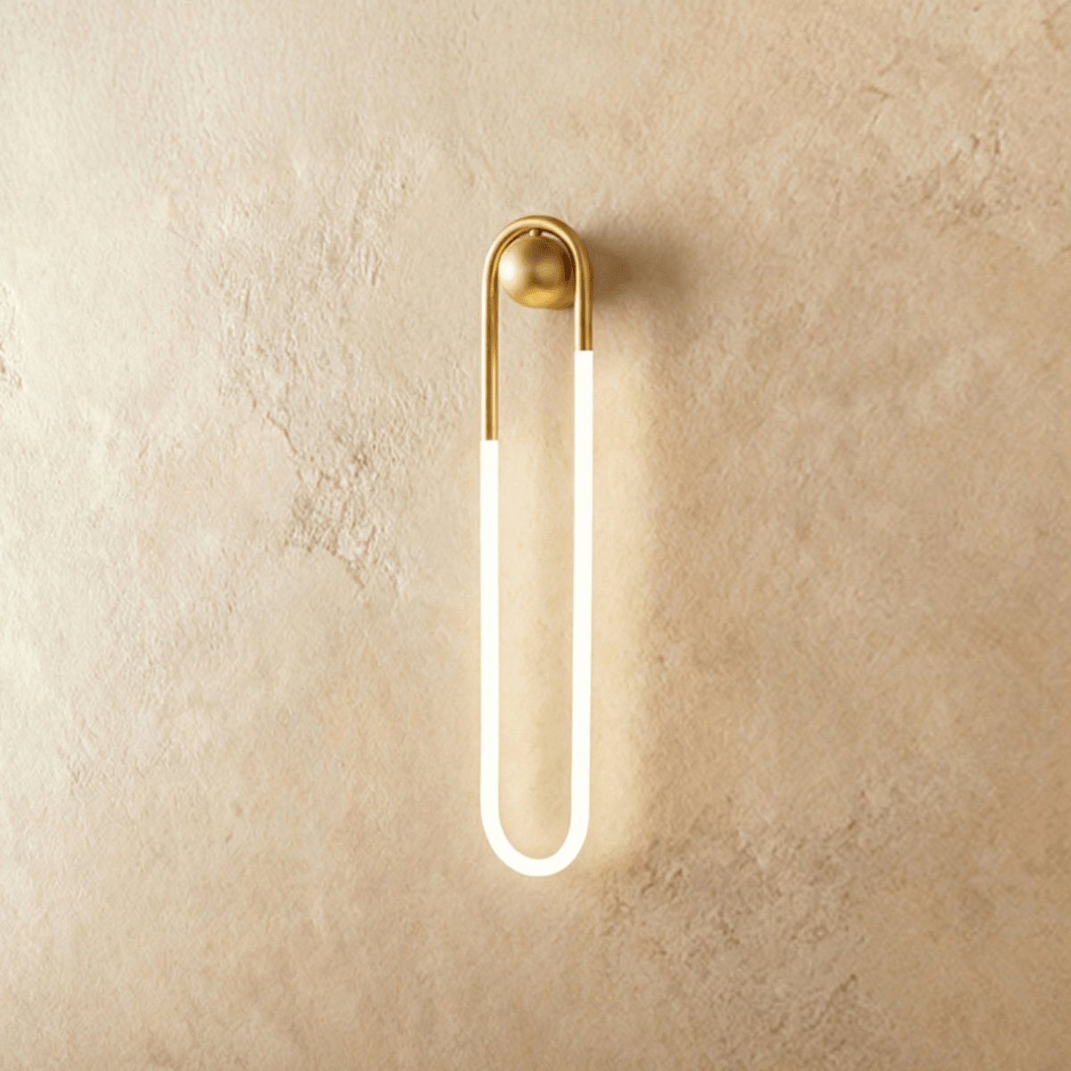 Clérachic slim U-shaped wall light with a brass arch and ball mount and a softly glowing frosted tube, on limewash plaster.