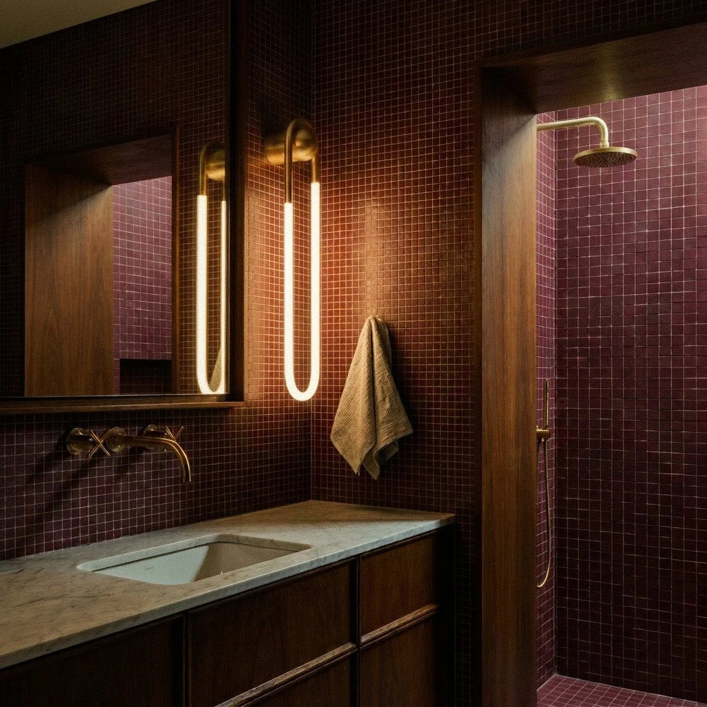 Clérachic brass U-shaped wall lights glowing beside a mirror in a bathroom of burgundy mosaic tile, with brass taps and a shower.